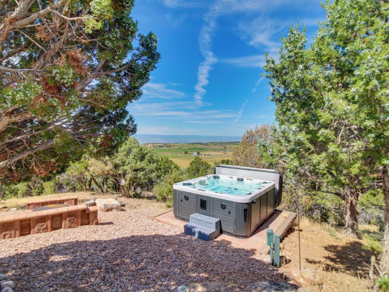 An outdoor setup surrounded by natural greenery and trees. In the background, a view of a vast valley stretches out under a blue sky with scattered clouds. The area includes a stone seating arrangement and is situated on a bed of gravel, blending into the landscape.