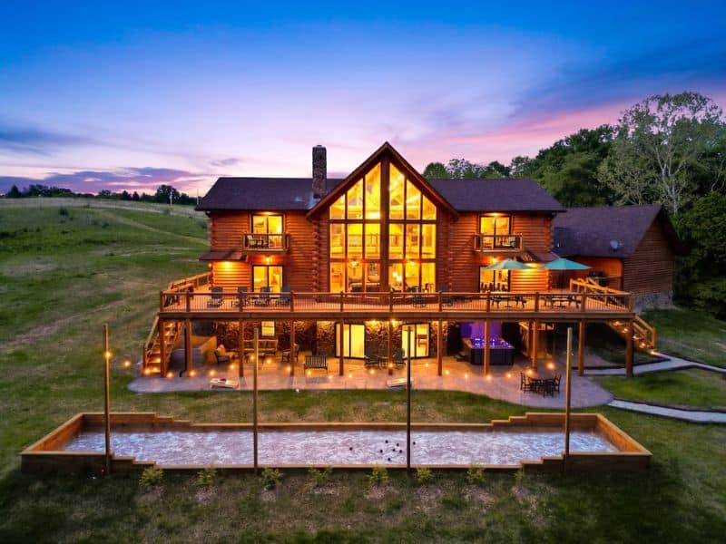 Dusk view of a grand wooden cabin with multiple levels of decks, illuminated windows, and an outdoor fire pit area in Hocking Hills, OH.