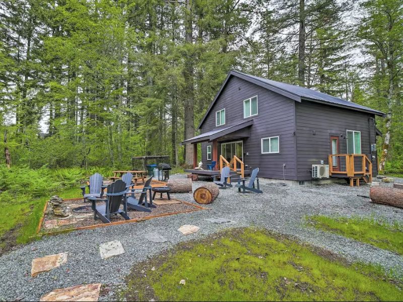 Black cabin exterior with outdoor hangout area including a fire pit, Adirondack chairs, and log seating. Part of a guide to the best Mt Rainier cabins