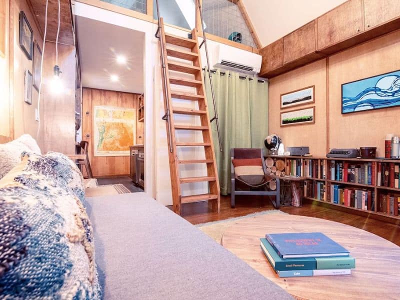 Interior of cozy A-frame cabin with ladder leading up to loft and living area with lots of books on book shelves