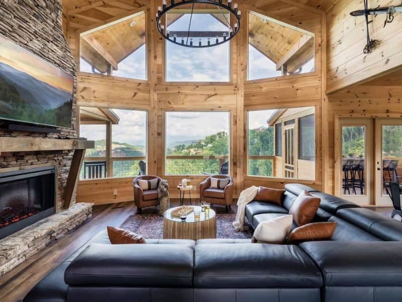 Modern-rustic living area in Gatlinburg cabin rental with floor to ceiling windows, stone fireplace, and large leather couch