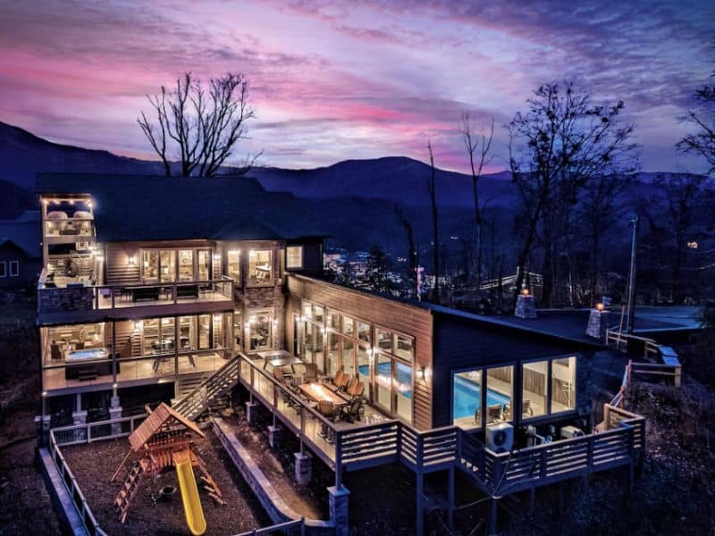 Modern luxury cabin lit up at dusk with large indoor pool and outdoor playground in Gatlinburg TN