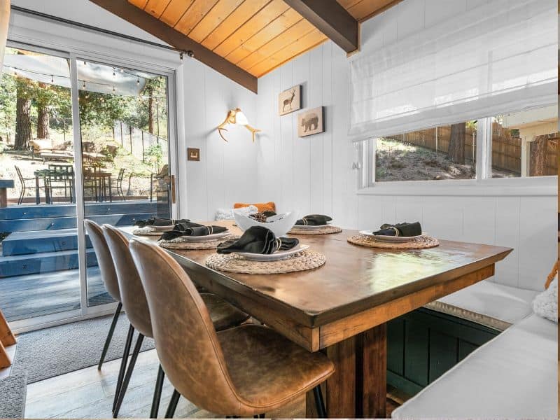 Charming dining area inside a cabin with a view of the deck and surrounding forest, featuring a rustic wooden table set for a gathering.