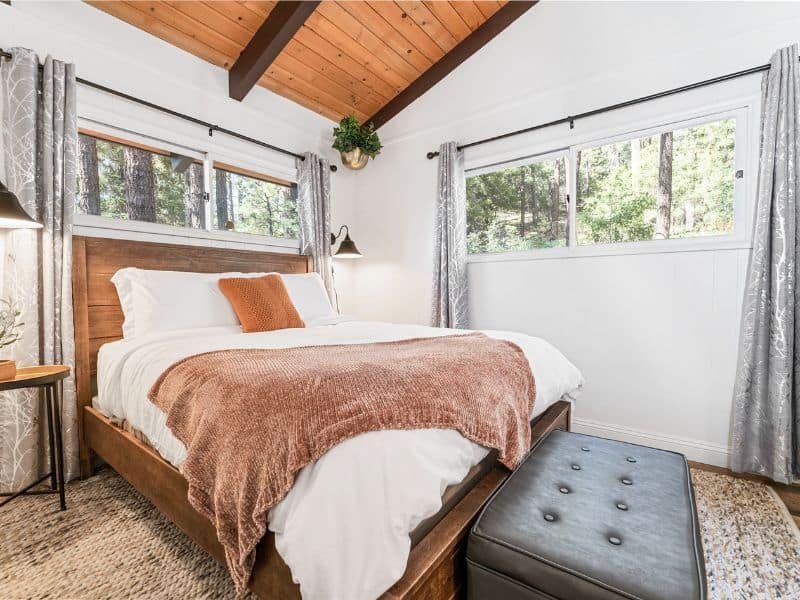 Cozy bedroom with a wood-framed bed, soft linens, and a view of pine trees in a Big Bear Vrbo cabin rental.