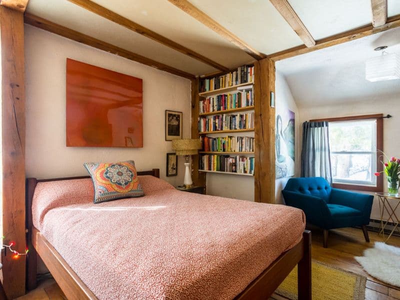 Cozy bedroom in a Catskills Vrbo rental with a full bookshelf, vibrant wall art, and a comfortable bed highlighted by natural light.
