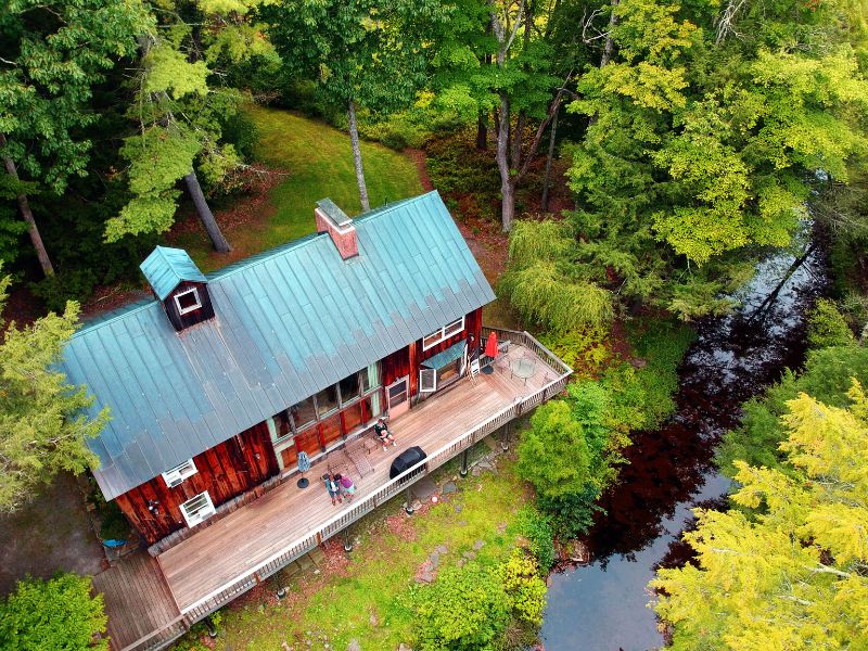 Aerial view of a two-story cozy Catskills cabin rental with a large deck, nestled among trees and alongside a calm stream.