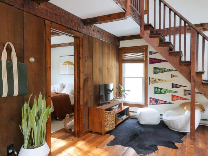 Rustic-chic lounge area of a Catskills rental cabin, with a wooden staircase, a collection of vintage pennants on the wall, and a snug TV nook leading to a bright bedroom.
