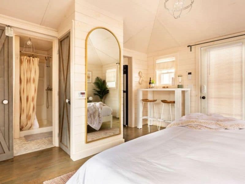 Bright and airy bedroom in a Catskills Vrbo rental, with a comfortable bed, reflective mirror, and a glimpse into the tiled bathroom.