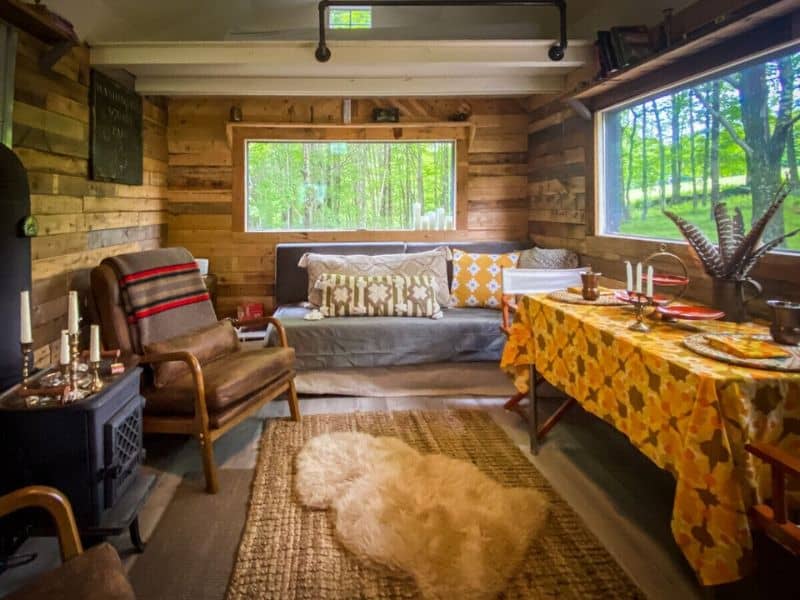 Inside a Catskills glamping cabin rental, a rustic living room with a wood-burning stove, leather armchair, and a dining area set against a backdrop of natural wood walls and a large window.