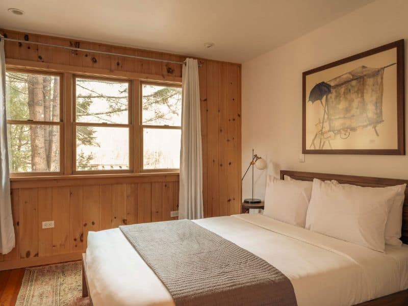 Simple yet elegant bedroom in a cozy cabin in the Catskills, with natural light streaming through large windows beside a comfy bed and artistic wall decor.