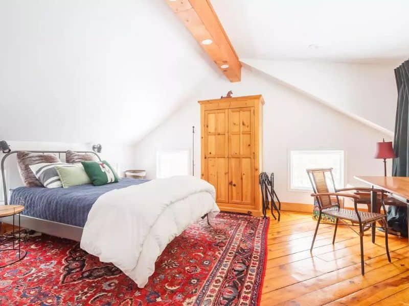 Bright and airy bedroom in a cozy cabin in the Catskills, with slanted ceilings, a wooden dresser, and a bed set against a patterned rug.