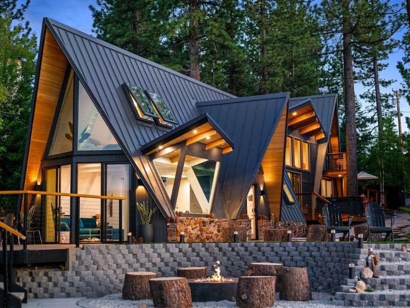 Architecturally distinctive A-frame lakefront cabin in Big Bear with illuminated interior, an outdoor fire pit, and seating area among the pines.