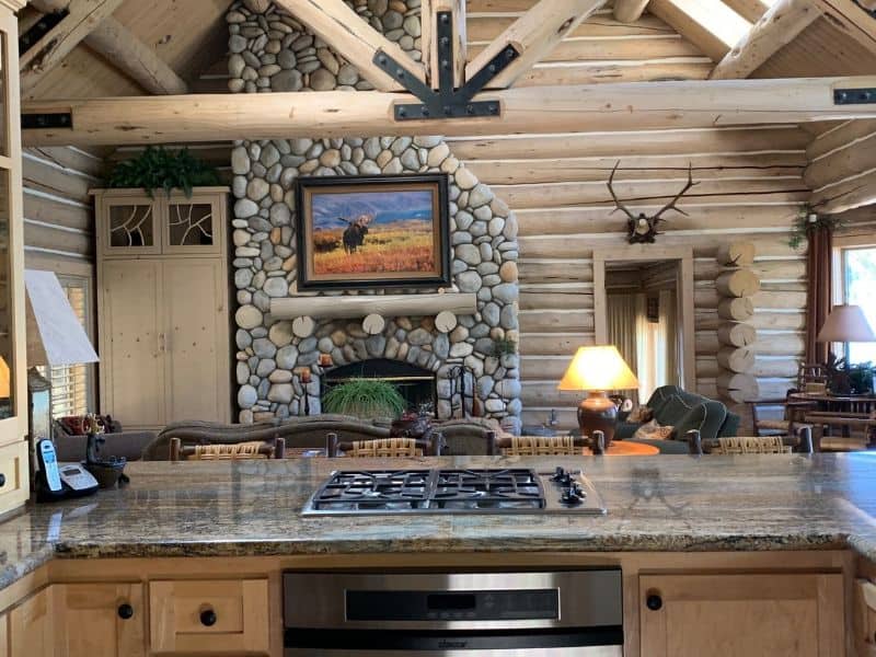 Open-plan kitchen looking into the living area of a Big Bear cabin rental with a stone fireplace and log cabin walls.