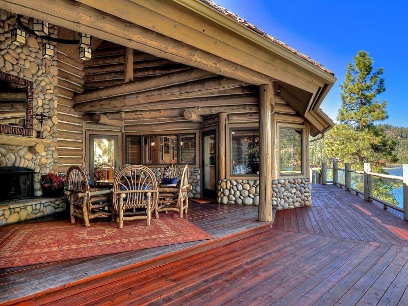 Rustic deck of a Big Bear lakefront cabin rental with log furniture and a large stone fireplace, with a view of the lake through the trees.