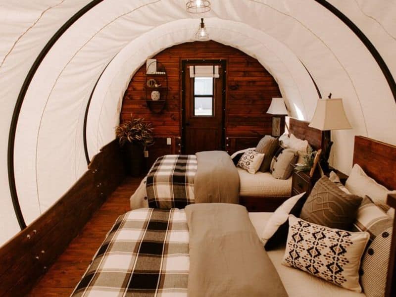 The interior of a covered wagon-style glamping unit featuring two neatly made beds with plaid bedding and decorative pillows. Covered wagon glamping at Grand Canyon Glamping Resort.