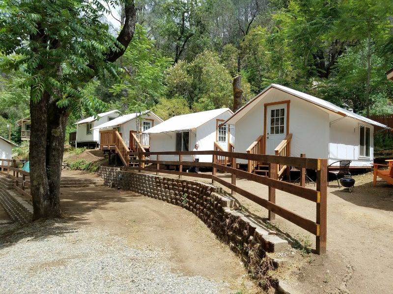 Canvas tent cabins on wooden platforms line a gravel path in a shaded grove at Indian Flat Campground, offering basic glamping near Yosemite.