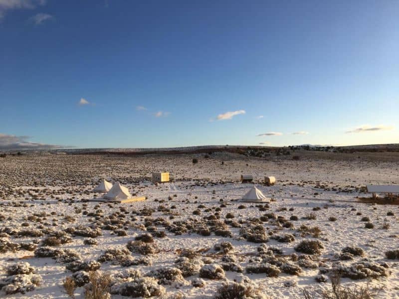 Glamping tents and huts scattered across a snowy landscape near the Grand Canyon, with clear blue skies and sparse vegetation.