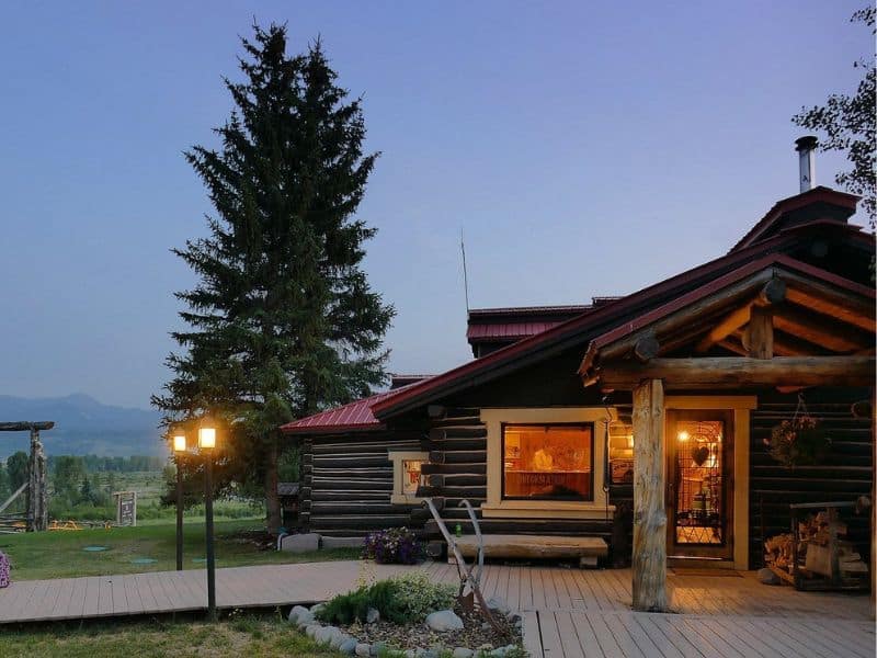 Exterior of the log cabin-style Heart Six Guest Ranch at twilight with lit lanterns and the silhouette of Grand Teton mountains in the distance.