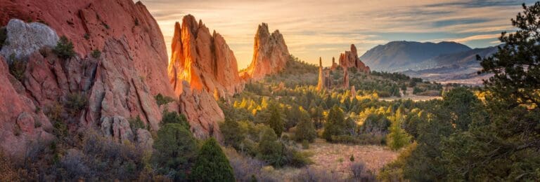 Red rock formations and lush greenery in Garden of the Gods, Colorado Springs, with the sun setting behind the mountains in the background. A great stop on a Colorado Utah road trip.